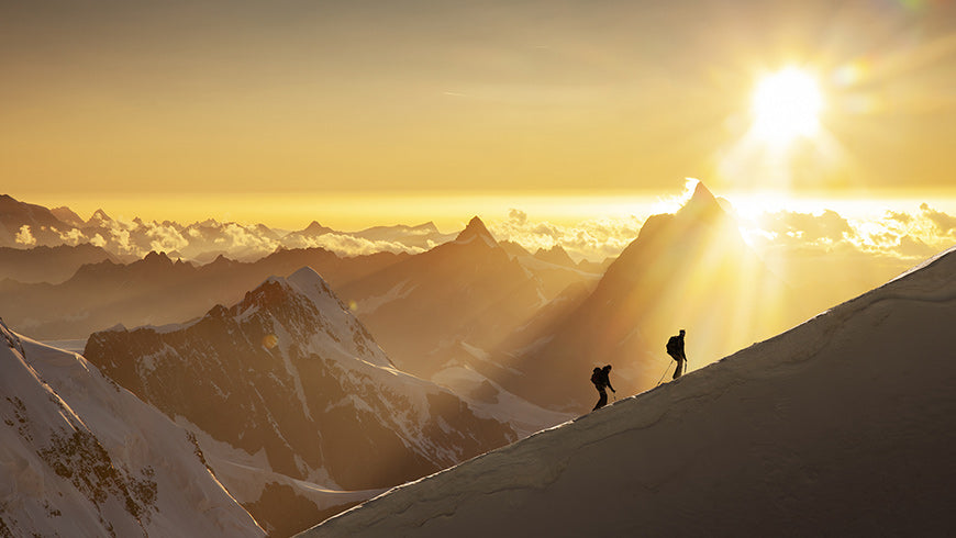 Two people hiking on a snowy mountain ridge with a golden sunset.